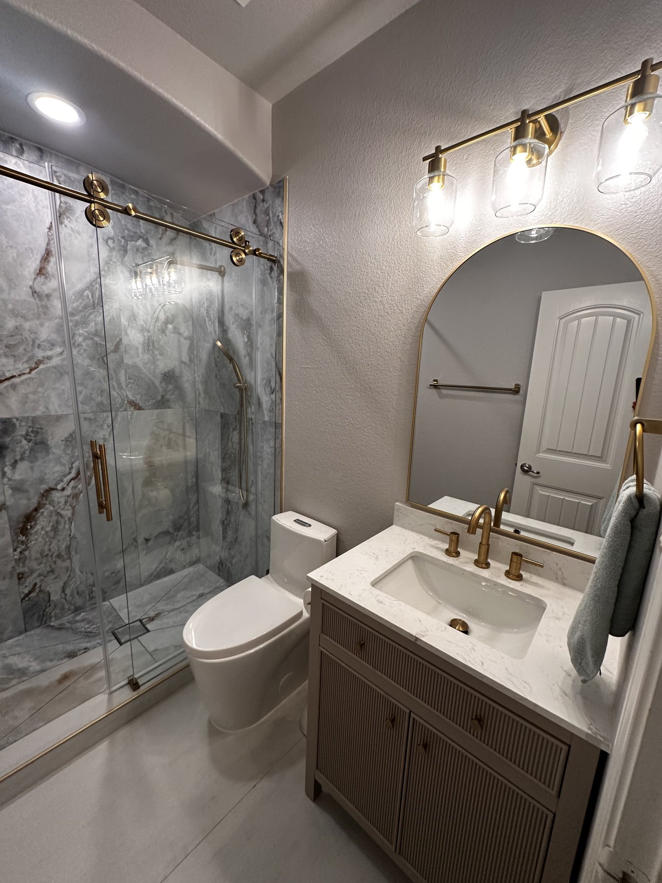 Modern powder bath with frameless sliding glass door, marbled slab, arched brass mirror, and fluted cabinetry — Forest Hills, Dallas TX